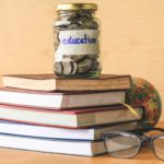 Coins in glass jar with education label, books,glasses and globe on wooden table. Financial concept.